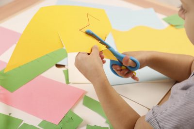 Little girl cutting color paper with scissors at table, closeup